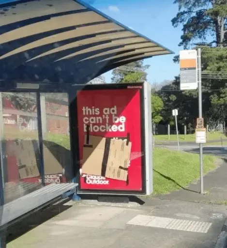 An advertising displayed on the wall of a bus stop. It reads "This ad can't be blocked". The rest of the ad is covered with a piece of cardboard. 